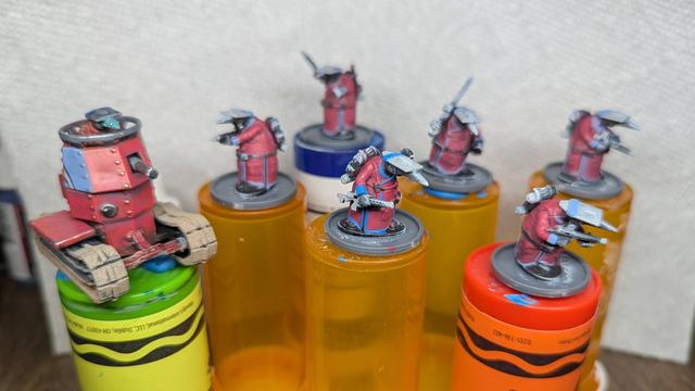 A group of partially painted plastic miniatures.  They are soldiers wearing flat metal hats and wearing long red coats with blue piping.  A one-person tank is part of the squad as well.  The soldiers are all Quar, a type of humanoid, anteatery, muppety thing.