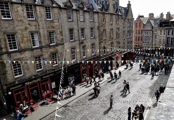Split-view of two cobblestone streets lively with pedestrians in a historic European city. The left image features a row of aged stone buildings with ground floor businesses, including a Mexican restaurant with red outdoor seating under colorful flag bunting. The right image captures a wider street scene bustling with people, lined with various shops under a series of multicolored flags stretched across from building to building, and a notable red and orange building visible in the background.