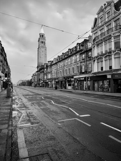 Black and white photograph of an urban street scene featuring a prominent stone tower with a pointed roof at the end of a street lined with four-story buildings. The road is marked with tram tracks and zebra crossings, and there is minimal traffic. Overhead, multiple wires run across the street. The image captures an overcast sky and wet pavement, suggesting recent rain.