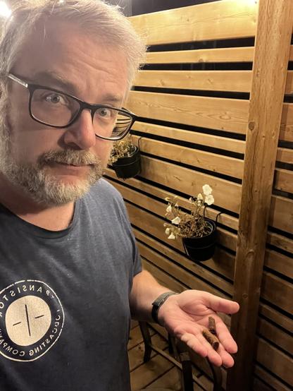 A person with glasses is holding a small object in their hand while standing in front of a wooden slatted wall with hanging flowerpots.