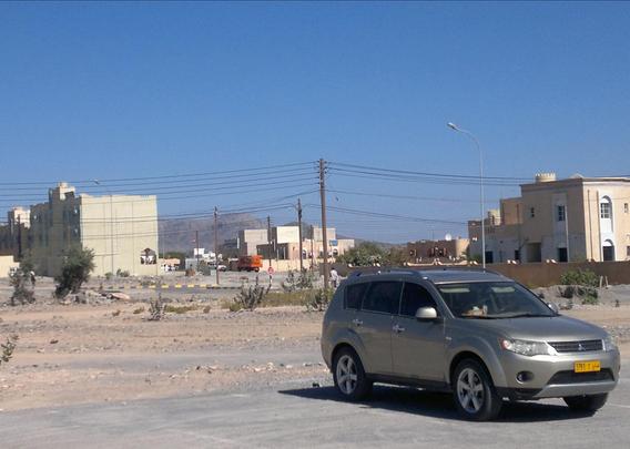 A silver SUV is parked on a dusty road with buildings in the background. The sky is clear and blue, and there are utility poles and some vegetation around.

Ein silberner SUV ist auf einer staubigen Straße mit Gebäuden im Hintergrund geparkt. Der Himmel ist klar und blau, und es gibt Strommasten und etwas Vegetation.