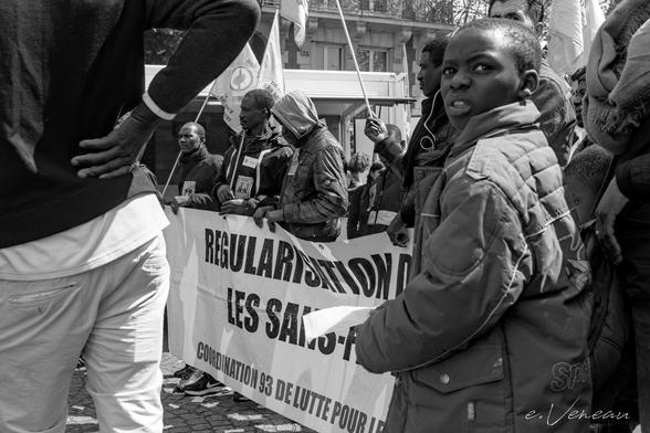 A group of workers join the march with a banner demanding their regularisation. In the foreground a child, face turned towards the photographer.