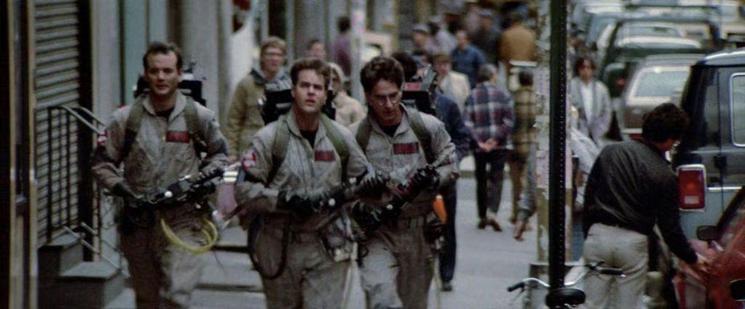 Three men in utility jumpsuits wear backpacks and hold gun-like items as they stroll down the street