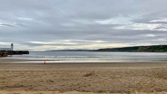 Scarborough Beach in England features a wide stretch of damp sand leading to calm, shallow waters under a cloudy, overcast sky. A lighthouse and pier are positioned to the left, marking the edge of the harbor, while a distant headland covered in green slopes extends along the right side of the bay. The tide is low, leaving wet sand patterns and a single orange cone near the shoreline. The sea is still, with faint ripples reflecting the muted light, and the horizon blends into the gray sky with only a few subtle breaks in the clouds.