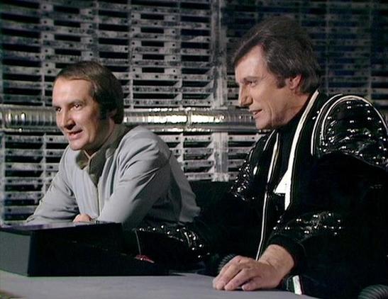 Claude 3.7 describes the image as: "The image shows a scene from a classic science fiction television series from the late 1970s/early 1980s. It depicts two characters in what appears to be a futuristic control room or spacecraft setting, with technical equipment and computer banks visible in the background.

On the left is a person wearing a light gray outfit, while on the right is someone dressed in a distinctive black leather or vinyl costume with silver accents. Both appear to be seated at some kind of control console. The setting has the characteristic visual aesthetic of British sci-fi productions of that era, with industrial-looking set design featuring computer banks and technical elements.

The scene appears to be taking place on the flight deck of a spacecraft, which was a common setting in this particular series."

Blakes7Bot, version 3.9.33.