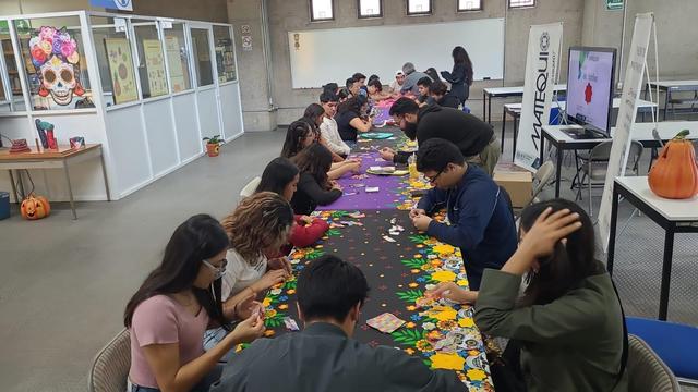 Foto de una mesa larga decorada con manteles alusivos a día de muertos. Varios jóvenes están sentados a la mesa haciendo figuras de papiroflexia.