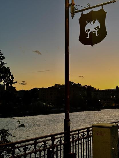 A waterfront scene featuring a decorative sign on a lamppost, silhouetted against a colorful sunset. The river is calm, and buildings are visible in the background. Green foliage is present in the foreground.