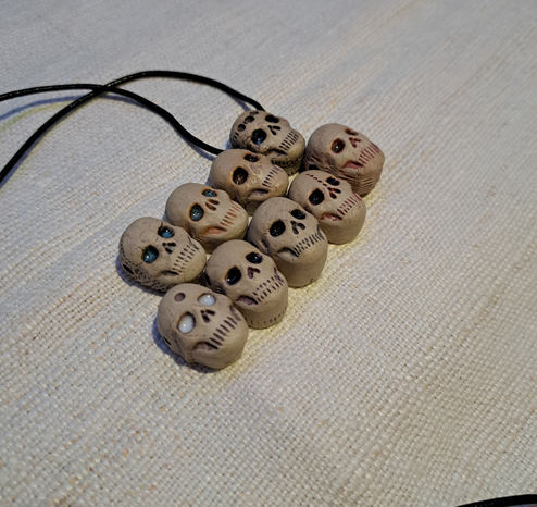 Nine stoneware clay pendants in the shape of skulls, all variations of the same, with differently coloured glazes in their eyes (though some look pretty much the same on the photo because of the lighting). One of them has a dark leather cord through it.