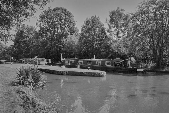 Black and white photo of a canal, showing a narrow boat being steered carefully into the lock to lie alongside one already there. The towpath is to the left, the lock in the centre, and trees behind.