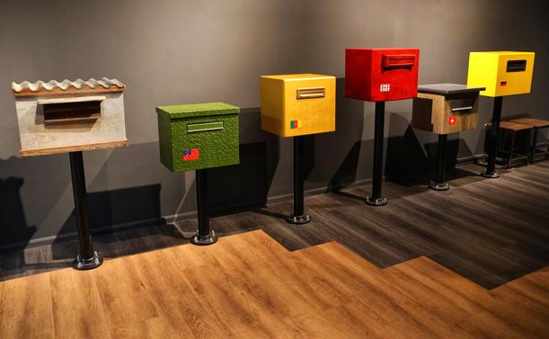 A row of five creatively designed, freestanding postal boxes displayed indoors on a wooden floor. From left to right:

1. A rustic-style box with a sloped, tiled roof and wooden sides, mounted on a black metal stand.
2. A green, textured box resembling artificial grass, featuring a small flag emblem.
3. A bright yellow box with a sleek, modern design and a small, multicoloured logo.
4. A bold red box with a minimalist design, displaying a white logo.
5. A yellow box with a black and red medical cross symbol, indicating a medical or emergency purpose.

Each box is elevated on a black metal stand, showcasing a variety of artistic and functional designs. The background features a neutral wall and geometric patterns on the floor.