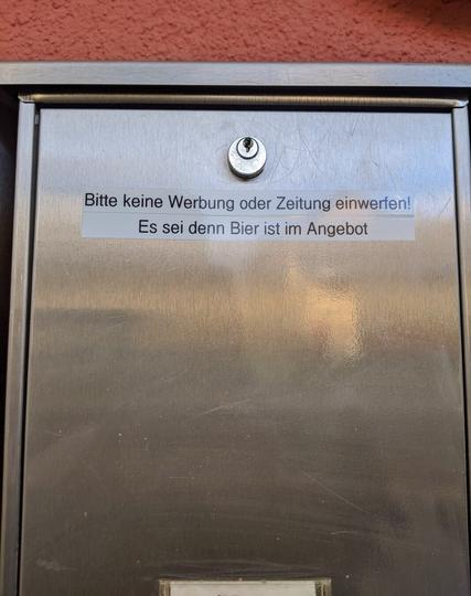 A close-up of a metallic, wall-mounted letterbox, likely located outside a building with a reddish-brown wall. The letterbox is made of brushed stainless steel and features a small lock at the top centre. A white label with black and blue text is affixed to the front of the box. The text, written in German, reads:

*"Bitte keine Werbung oder Zeitung einwerfen! Es sei denn Bier ist im Angebot"*

This translates to: "Please do not insert any advertising or newspapers! Unless there is beer on offer." The letterbox has a slot for mail on the top and a lock, and the surface shows minor signs of wear and reflections.