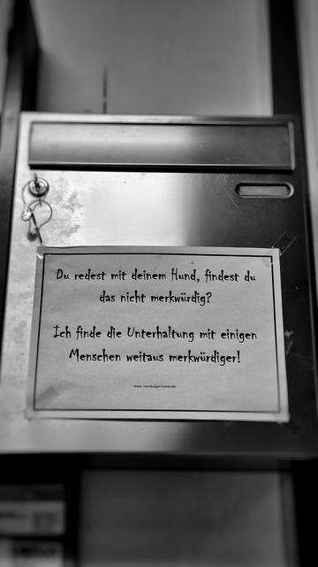 Schwarz-Weiß-Foto eines Briefkastens aus Edelstahl, aufgenommen von vorne unten nach oben. Oben ist die Klappe für den Einwurf zu sehen, darunter links das Schloss mit Schlüssel und rechts das unbeschriftete Sichtfenster für das Namensschild. Auf der Briefkastentür ist ein DIN-A4-Blatt quer angebracht mit der Aufschrift „Du redest mit deinem Hund, findest du das nicht merkwürdig? Ich finde die Unterhaltung mit einigen Menschen weitaus merkwürdiger!”