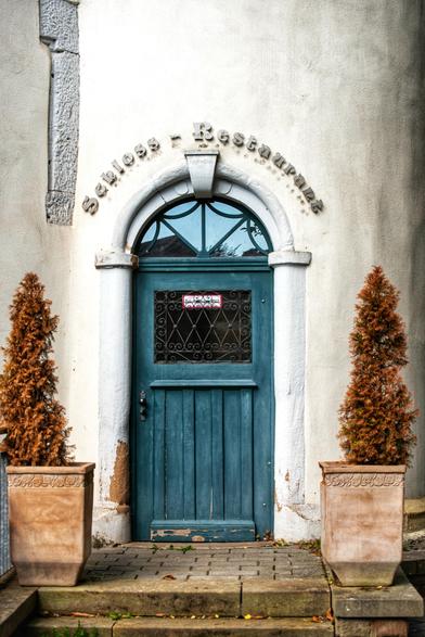Das Bild zeigt einen Eingang aus einer blauen Holztür mit einem Rundbogen, über dem in Metallbuchstaben der Name "Schloss - Restaurant" angebracht ist. Im Fenster der Tür ist ein Schild mit der Aufschrift 'Zuluftöffnung' angebracht. 
Die Tür ist in eine helle, verputzte Wand eingelassen und wird von zwei weißen Säulen eingerahmt. Links und rechts der Tür stehen zwei in Töpfen gepflanzte, vertrocknete Nadelbäume.