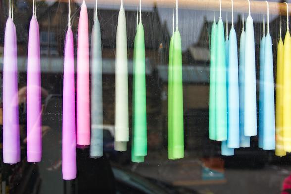 The photo shows a row of pastel-coloured candles hanging in a shop window. The candles are long and slender, suspended by their wicks, and arranged in a gentle gradient — from soft pinks and lilacs on the left to greens, blues, and a hint of yellow on the right. The glass reflects the street outside, with faint shapes of buildings and the photographer visible in the lower left, blending into the scene.