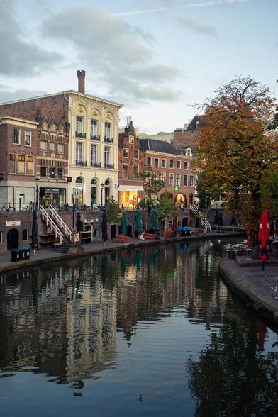 This image depicts a serene canal scene in an urban area. The canal, calm and reflective, runs through the center of the image, mirroring the buildings and trees along its banks. On the left side of the canal, there are several multi-story buildings with varied architectural styles. The building closest to the viewer is a light-colored structure with ornate details, including arched windows and balconies. Adjacent to it are other buildings made of brick, featuring large windows and some with small balconies.

The right side of the canal is lined with trees, their leaves displaying autumn colors of orange and yellow. There are outdoor seating areas with tables and chairs, some covered by umbrellas, suggesting the presence of cafes or restaurants. The sky above is overcast with a mix of light and dark clouds, casting a soft light over the scene. The overall atmosphere is tranquil and picturesque, capturing the charm of a canal-side urban setting.