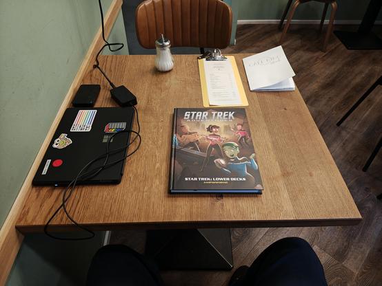 laptop zugeklappt links, mit einigen rainbow stickers, Star Trek Lower Decks Kampagnenband in der Mitte auf einem hölzernen Tisch