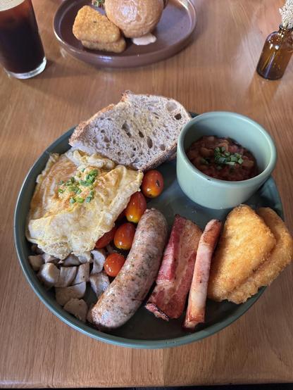 An overhead shot shows a full breakfast plate with eggs, bread, sausage, bacon, beans, tomatoes, hash browns, and mushrooms.