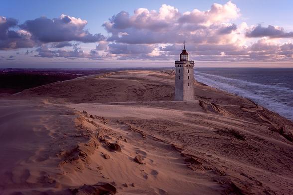 The Rubjerg Knude Fyr lighthouse stands on a sandy dune along the North Sea coast in northern Denmark. The tower is rectangular, made of light-colored stone, and topped with a red roof and lantern. The surrounding area consists of extensive sand dunes stretching to the horizon. The sky features a mix of clouds and blue, illuminated by the warm light of the setting sun.