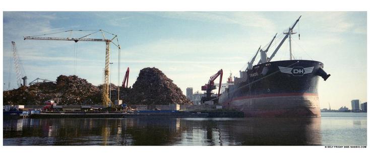 Colour panorama photograph of a huge Amsterdam (West) dockside pile of scrap iron. On the right is a large bulk carrier being loaded by several cranes on board and on the harbour quay. To the left is another lighter hoisting L crane unloading a smaller barge.