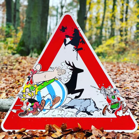 Das Bild zeigt ein Verkehrsschild, das in einem herbstlichen Wald aufgestellt wurde. Das dreieckige Schild hat den typischen roten Rand eines Warnschilds, aber anstatt einer herkömmlichen Warnung sieht man eine Szene mit den Comicfiguren Asterix und Obelix, die mit anderen Tieren von links nach rechts rennen. Im Hintergrund sieht man einen Wald mit braunen Herbstblättern, die den Boden bedecken.