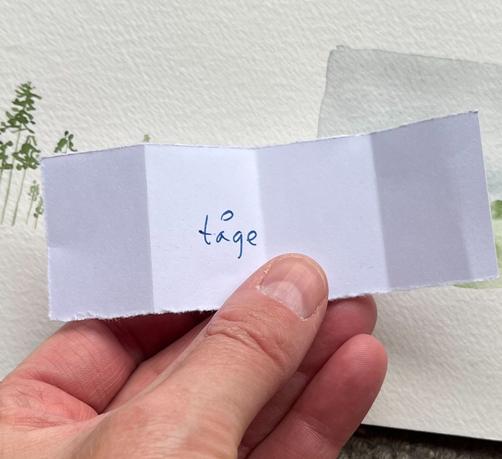 Foto af en hånd med en seddel, hvor der står tåge med håndskrift. Baggrunden er en maleblok med to forskellige motiver, som dog er primært skjult bag sedlen.