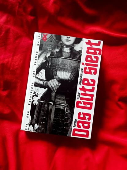 Foto des Buches, auf dessen Schwarz-Weiß-Cover Teile einer gerüsteten Frau (Jeanne d’Arc) zu sehen sind, die mit der rechten Hand ein vor sich aufrecht hingestelltes Schwert hält; oberhalb der Wangenknochen und etwa ab den Knien nach unten ist das Bild beschnitten. Hinter ihr sind weiße Lilien und ein Kirchenbau mit einer Fensterrosette zu sehen. Das Buch liegt auf einem roten, Falten werfenden Stoff, der die Farbe der Schrift auf dem Cover aufnimmt.