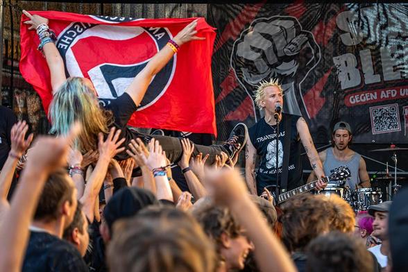 Das Foto zeigt eine Szene des Konzerts: Ein Mensch mit Antifafahne beim Stagediving, dahinter die Bühne mit den Musikern