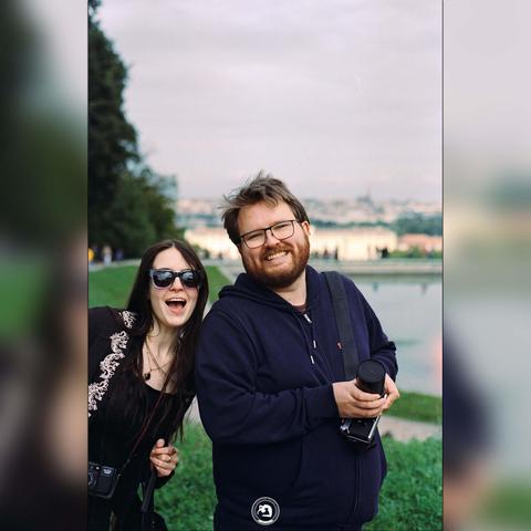 A colour photo of a woman with dark hair and sun glasses and a bearded man with glasses posing in front of Schönbrunn castle, Vienna, looking towards the camera. The man is holding a camera in his hand.