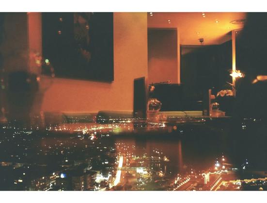 A dimly lit lounge with cozy seating reflects a vibrant cityscape at night through large windows. Warm amber tones evoke an intimate, urban atmosphere.