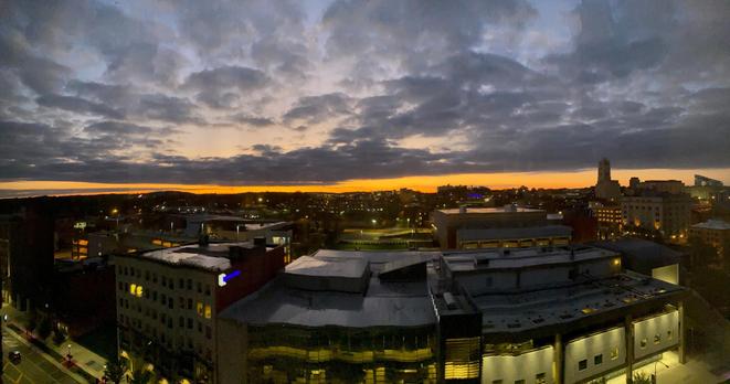 Color image of pre-dawn downtown Akron, Summit County, Ohio, USA. Image shows the brightening sky under a layer of clouds, the sun still beyond the horizon beginning to light the day, and orange and yellow band of light illuminates the far horizon, the city below still asleep.