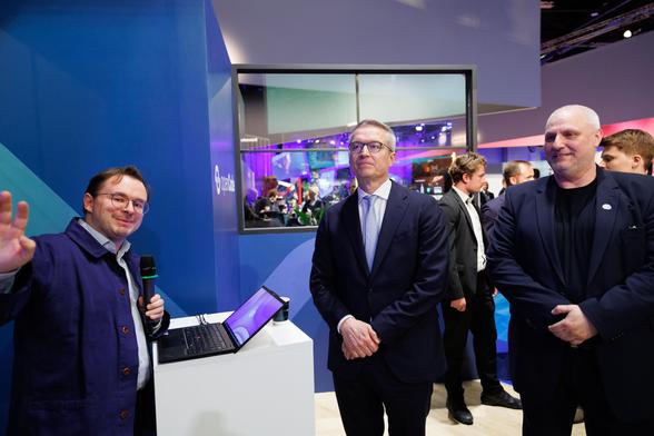 Alexander Smolianitski steht vor einem Pult, auf dem ein Laptop steht. Er hält ein Mikro in der linken Hand und zeigt mit der rechten in der Kamera. Karsten Wildberger und Alexander Pockrandt stehen rechts neben ihm.