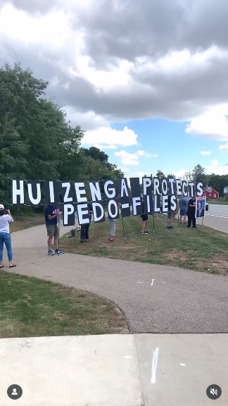 People standing on a public sidewalk holding a big sign that says "Huizenga protects pedo-files." It's partly cloudy with pretty trees in the background. Great place to say release the files.