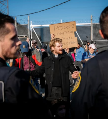 A young white priest with blonde, wavy hair, wearing black clothes and clerical collar, stands peacefully in front of a group of ICE agents (I think) just talking to them. Behind him are other protesters and some cameras that may be from local media. The illegal chain link fence at the Broadview ICE facility, which ICE put up and which the local fire marshall says is a danger, is visible in the background.