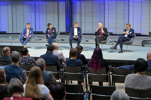 Vier Männer und eine Frau sitzen auf Stühlen auf einer Bühne und diskutieren.