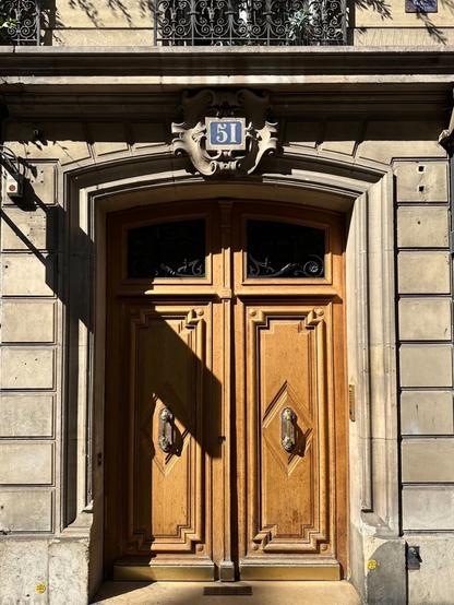 an ornately carved wooden double door in a stone building. Above the arched doorway, a blue plaque reads 51