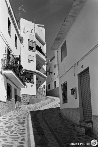 Black and white photo of a narrow, winding street in Guájar-Fondón, lined with white buildings. Patterned paving and balconies with railings catch the light. Shadows reveal bright sunlight.