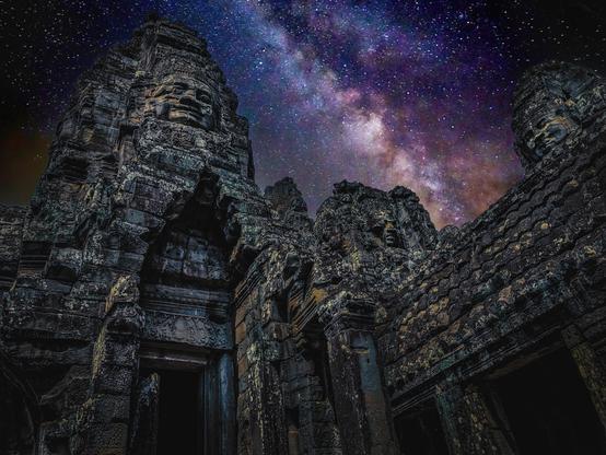 Night photograph of Bayon Temple in Cambodia, showing several stone towers carved with giant serene faces, silhouetted against a vivid Milky Way sky of purples, blues, and pinks.