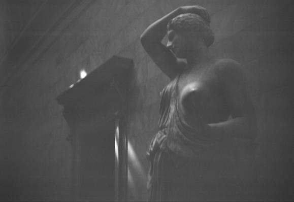 The monochrome photograph provides a low angle view of Roman marble copy of a Greek bronze sculpture of a female figure, right arm raised to its head, its torso half covered by its dress.