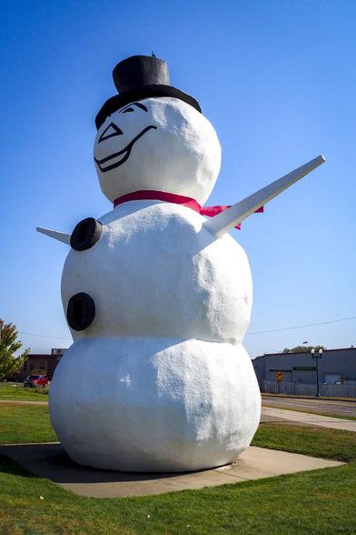 A giant snowman statue wearing a red scarf and black top hat along the side of a road