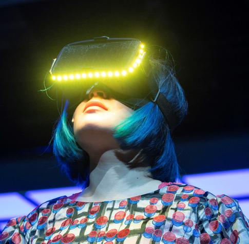 Eine Frau mit türkis-blauem Bob-Haarschnitt trägt eine große, schwarze VR-Brille mit leuchtendem LED-Rand. Ihr Blick geht leicht nach oben, die Lippen geschlossen. Ihre Kleidung ist bunt gemustert. Das Bild zeigt staunende Immersion und digitales Erleben.