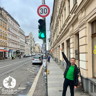 Eine Straße mit mehrstöckigen Altbauten auf beiden Seiten. Ein Ampelmast zeigt grünes Licht, darüber ein rundes Verkehrsschild mit der Zahl '30' in rotem Kreis. Am rechten Gehweg steht eine Person in schwarzer Jacke und grünem T-Shirt, die mit dem rechten Arm nach oben zeigt. Im Hintergrund sind mehrere geparkte Autos und weitere Fußgänger zu sehen. Unten links ist ein weißes Logo mit einem Löwen und dem Schriftzug 'Ökolöwe'.