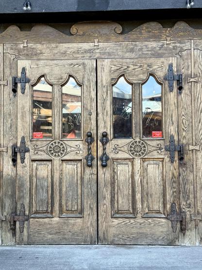 Photo of a set of wooden doors with black metal door handles and hinges.  The doors have four panels with the upper two having glass inserts.  Between the door panels are decorative carvings in a flower pattern.