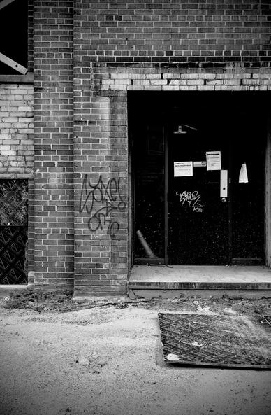 Brick building under construction. Windows to the left have cross braced wood support and the windows are removed. To the right is an alcove with a door and construction notices.