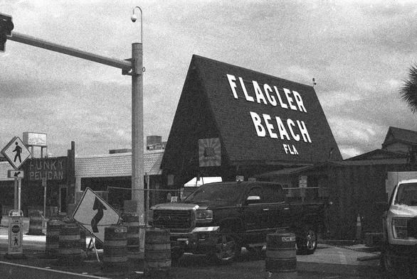 A building is surrounded by fences and barrels. A sign on the low part of the building reads “Funky Pelican”. There is a high pitched roof at the right end of the building, perpendicular to the rest of the building, and huge letters attached to the roof that read “Flagler Beach FLA”.