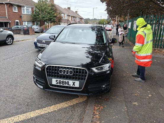 A very badly-parked car on yellow zigzag lines. A man in a hi-vis cost is pointing down the pavement as a woman approaches carrying a child
