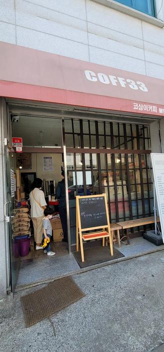 A storefront with a pink awning that says "COFF33" in white letters is captured in an eye-level shot. A small chalkboard menu is displayed outside the entrance. People are visible inside the store.