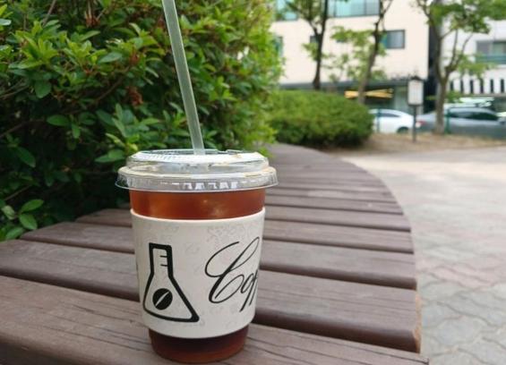 A coffee cup with a straw rests on a wooden bench outdoors.