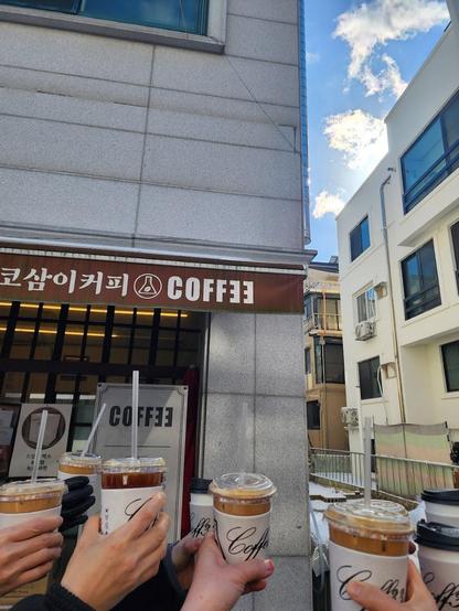 A group of people holding up iced coffee drinks in front of a coffee shop.