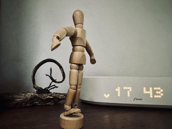 Une freebox blanche qui indique 17:43, à côté une sorte de sculpture en algues et en écorces, au premier plan un mannequin en bois miniature, pied levé et bras en mouvement.