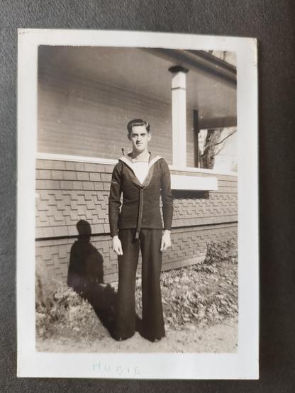 A tall lanky man in a navy uniform back in the 1940s. The photo is black and white and he's standing in front of a house. His hair is done in a very common 1940s style for men, slicked back and short sides.